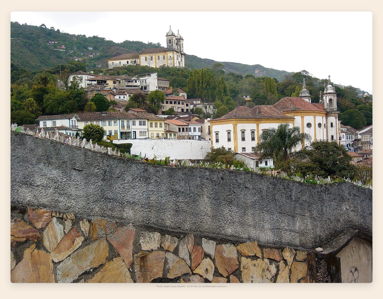 Ouro Preto's pastel townhouses and twin-towered baroque church climb a green hillside above a stone wall