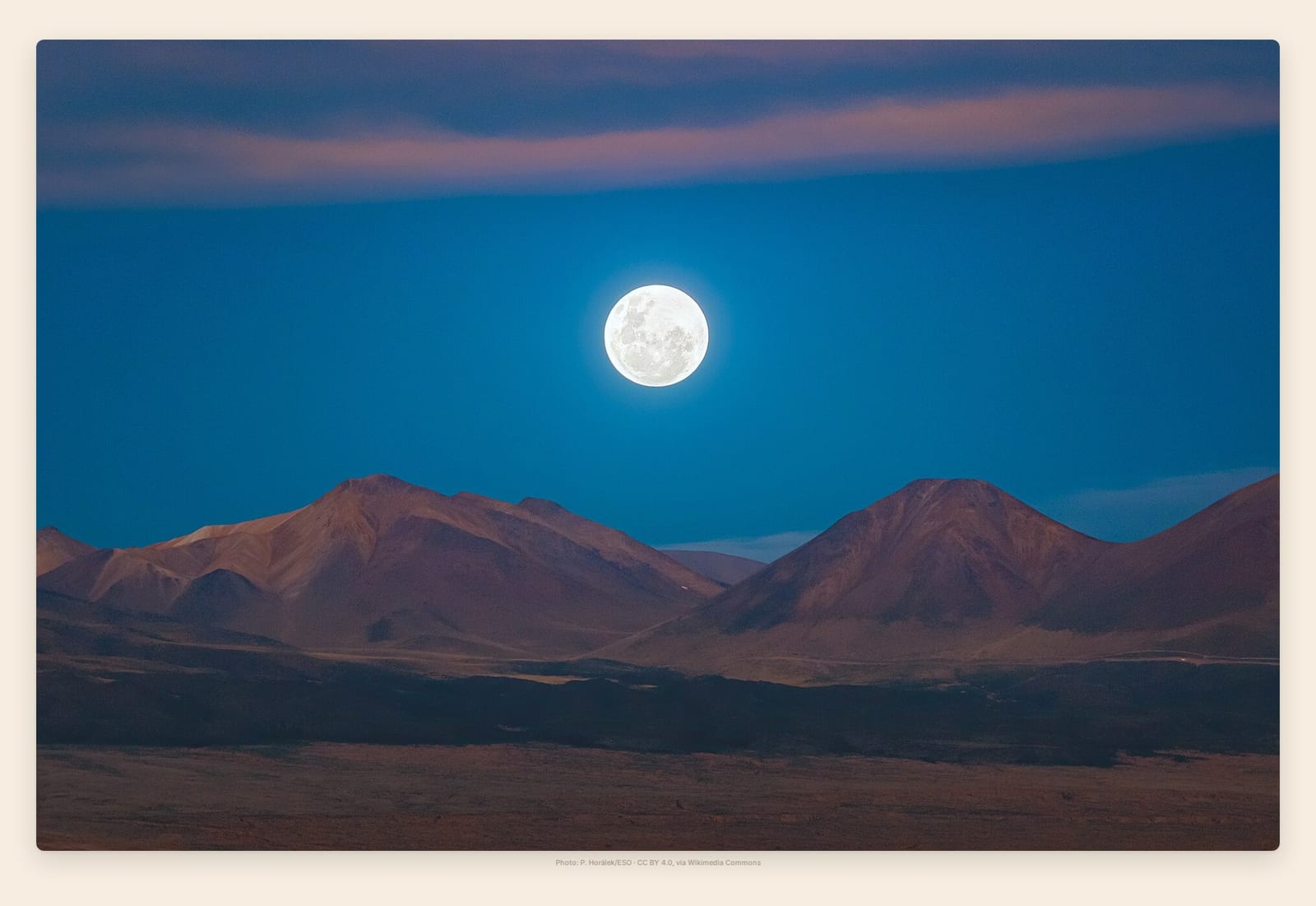 A huge bright full moon dominates a nearly starless twilight sky above a mountain range