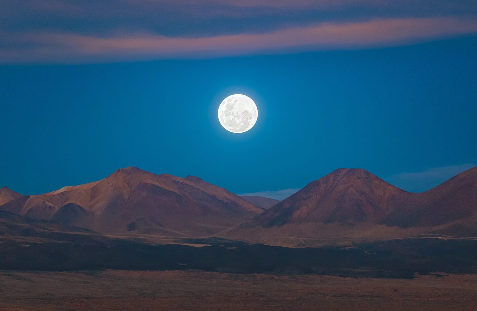 A huge bright full moon dominates a nearly starless twilight sky above a mountain range