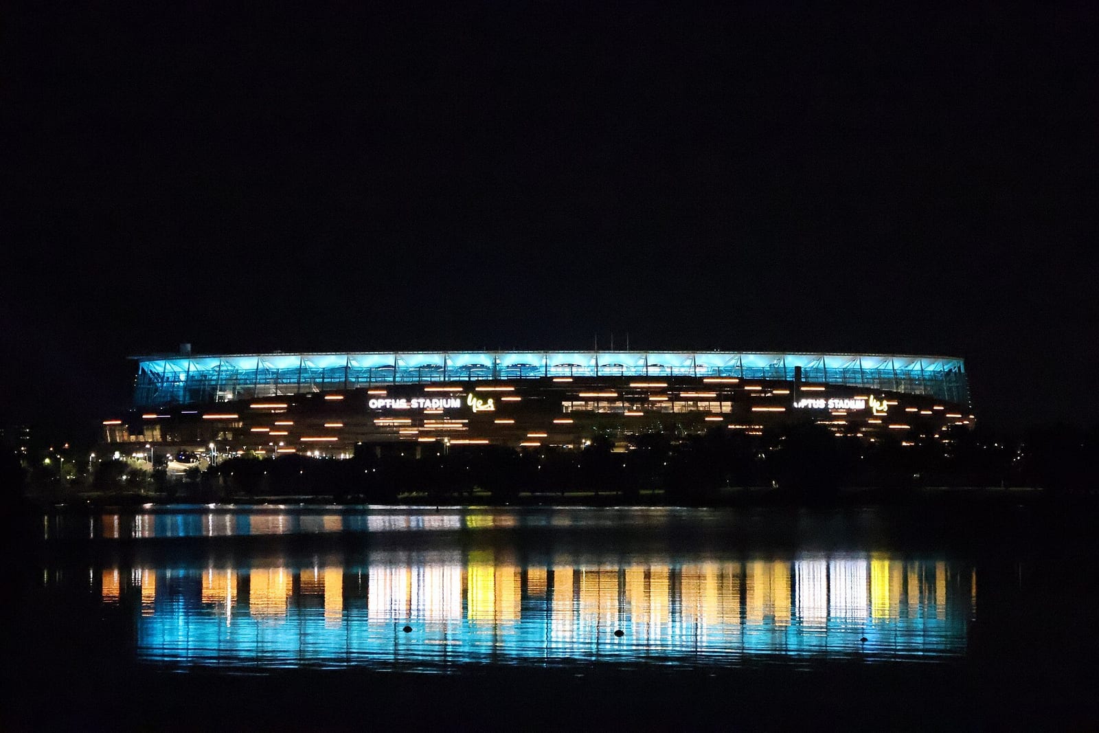 Optus Stadium in Perth glowing at night, its lit roof and windows reflected in the river