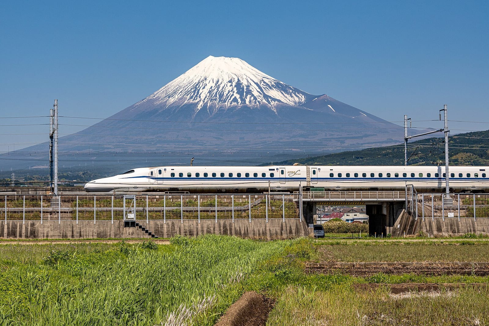 A white N700S Shinkansen crosses farmland on the Tokyo-Kyoto line with snow-capped Mount Fuji behind it